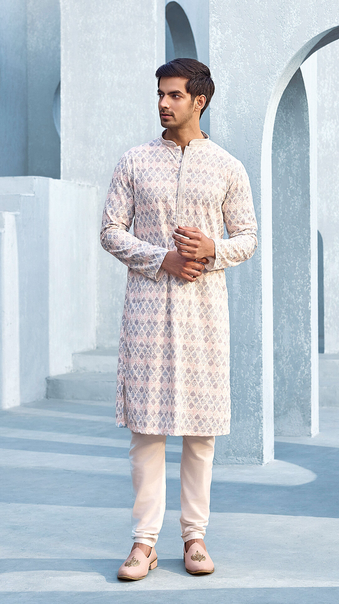 Man wearing a pastel printed kurta pajama set with traditional mojaris, posing against a modern architectural backdrop in soft blue tones